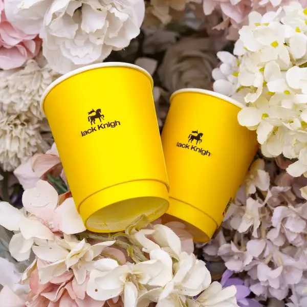 Copos de papel amarelos personalizados impressos com o logo do Cavaleiro Negro colocados entre as flores